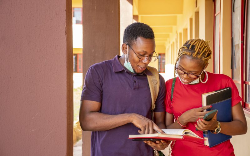 Two young African students wearing facemasks and holding books and phones at a campus